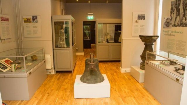 A museum room with glass display cases containing historical artifacts, documents, and a large bell in the center on a white pedestal, all on a wooden floor.