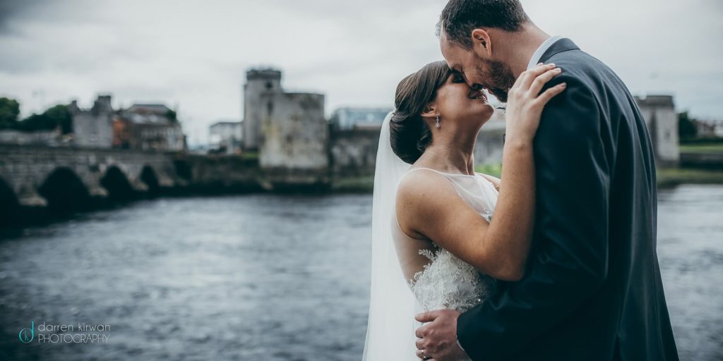 A bride and groom embrace and smile at each other by a river, with stone buildings and a bridge in the background on a cloudy day.