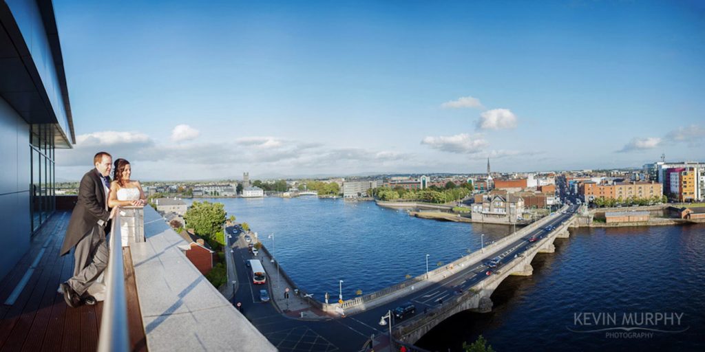 A couple stands on a balcony overlooking a wide river, bridge, and cityscape under a partly cloudy sky.