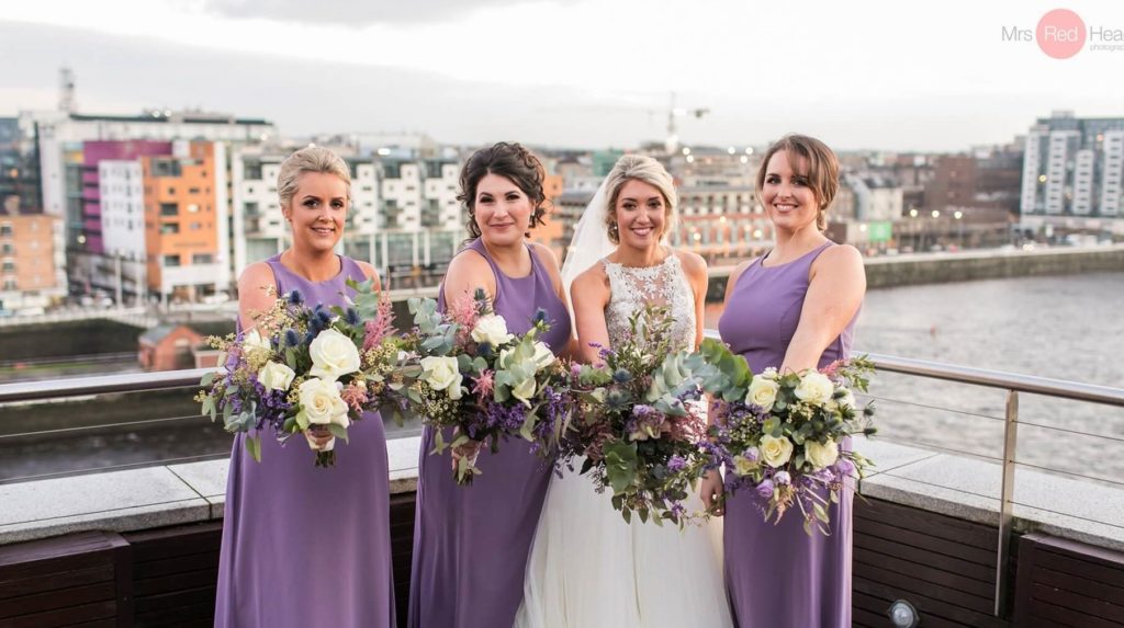 Four women pose on a rooftop by a river; three wear matching lavender dresses holding bouquets, one wears a white wedding gown and veil.