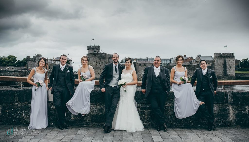 A wedding party of four women in white dresses and four men in dark suits pose together outdoors by a stone wall, with a large historic castle in the background under cloudy skies.
