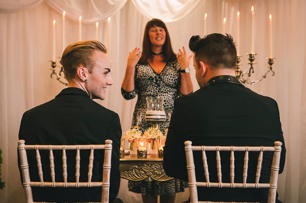Two men in suits sit facing a woman standing and speaking in front of them at a decorated indoor ceremony with candles and draped fabric.