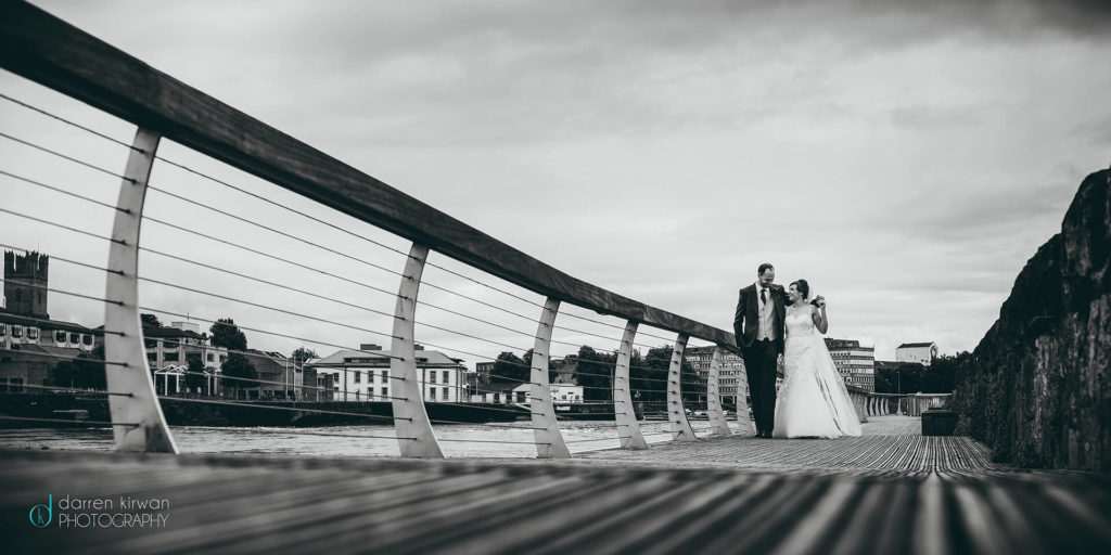 A bride and groom walk together on a riverside boardwalk, with buildings and trees visible in the background under a cloudy sky.