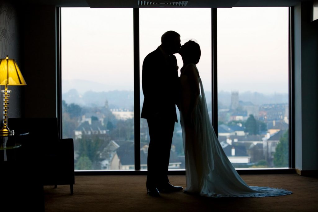Silhouette of a couple standing closely together by a large window, with a cityscape visible in the background at dusk.