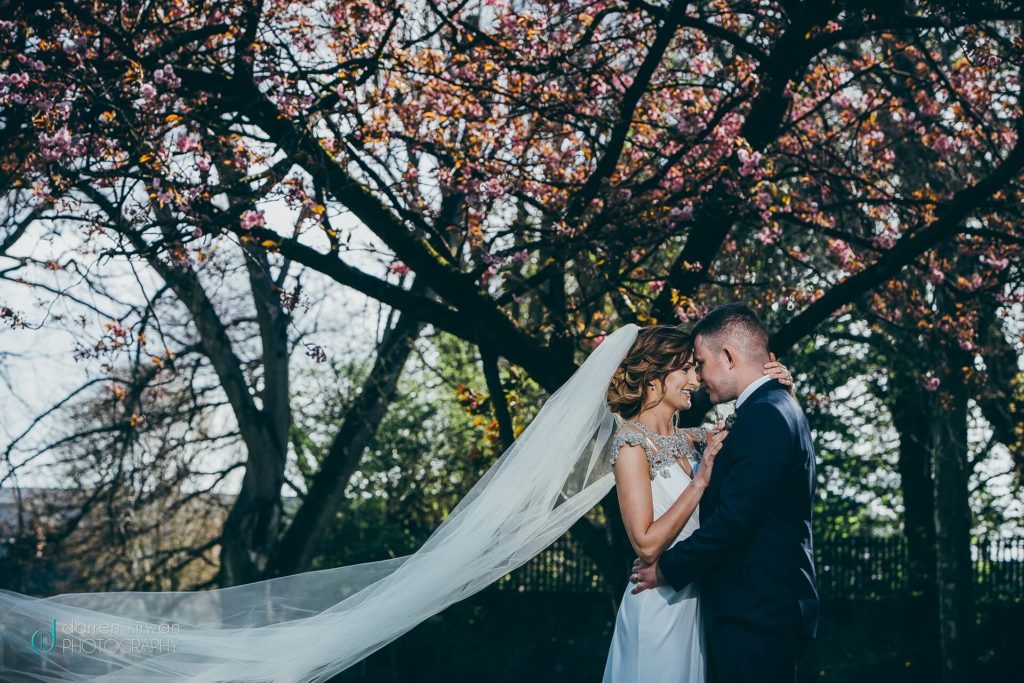 A bride and groom stand close together under a blooming tree, with the bride’s veil flowing behind her on a sunny day.