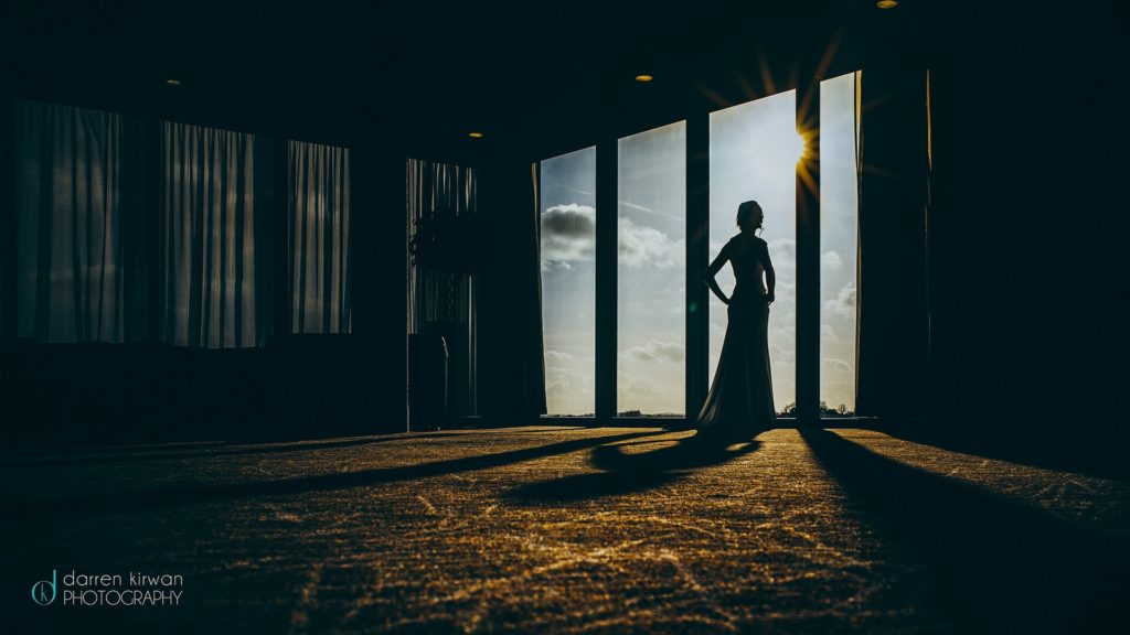 A silhouetted person stands near tall windows with sunlight streaming in, casting long shadows across the floor in a dimly lit room.