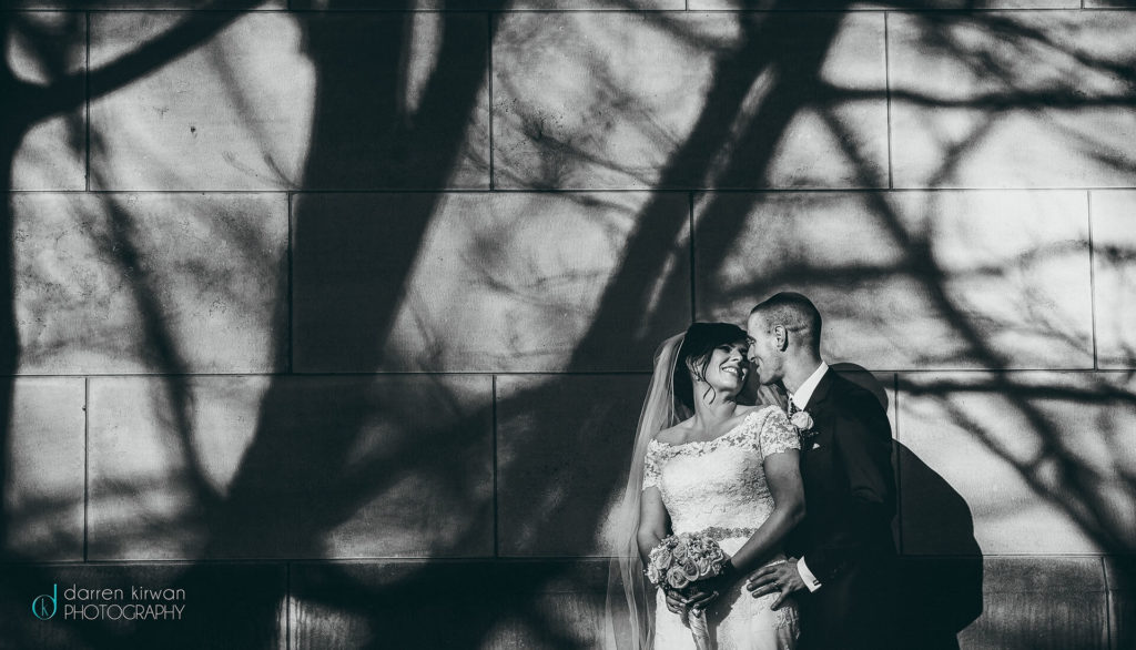 A bride and groom stand close together against a sunlit wall with dramatic tree branch shadows. The bride holds a bouquet and smiles while the groom stands behind her.