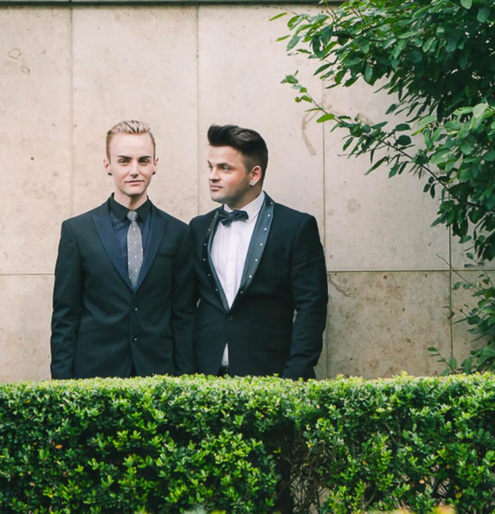 Two people in formal suits stand side by side outdoors in front of a stone wall and green hedge, with leafy branches on the right.