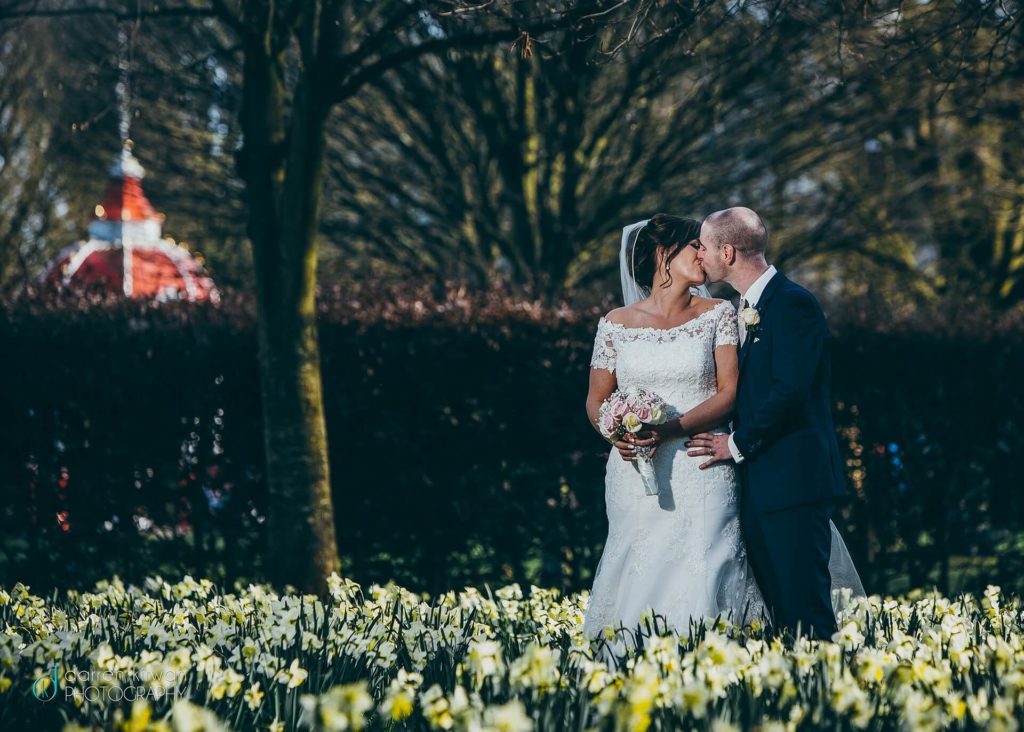 A bride and groom stand close together among blooming daffodils, with the groom kissing the bride’s cheek. Trees and a red-roofed structure are visible in the background.
