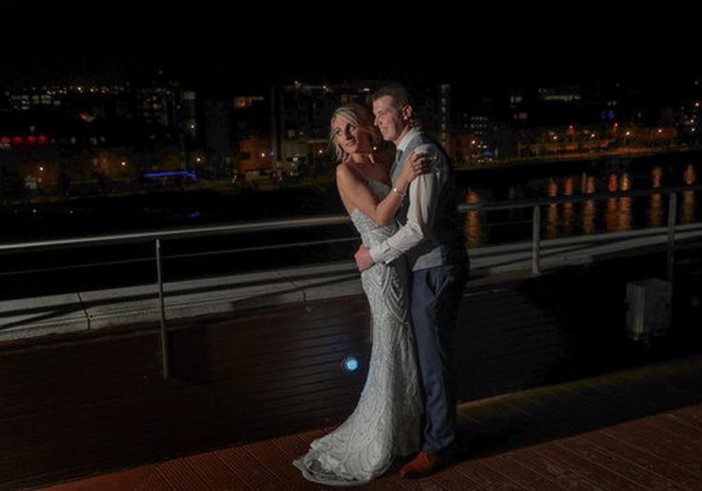 A bride in a white gown and a groom in a gray vest embrace and smile on an outdoor deck at night with city lights in the background.