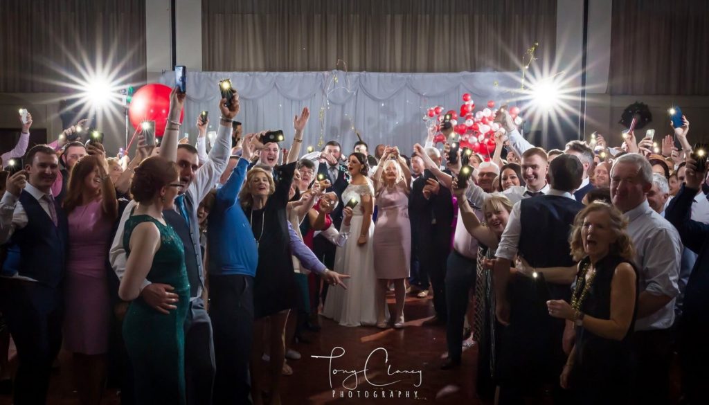 A large group of people dressed formally stand together indoors, raising glasses and smiling at the camera, with bright lights illuminating the scene.