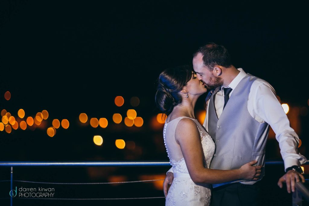 A bride and groom kiss at night on a balcony, with blurred city lights in the background.