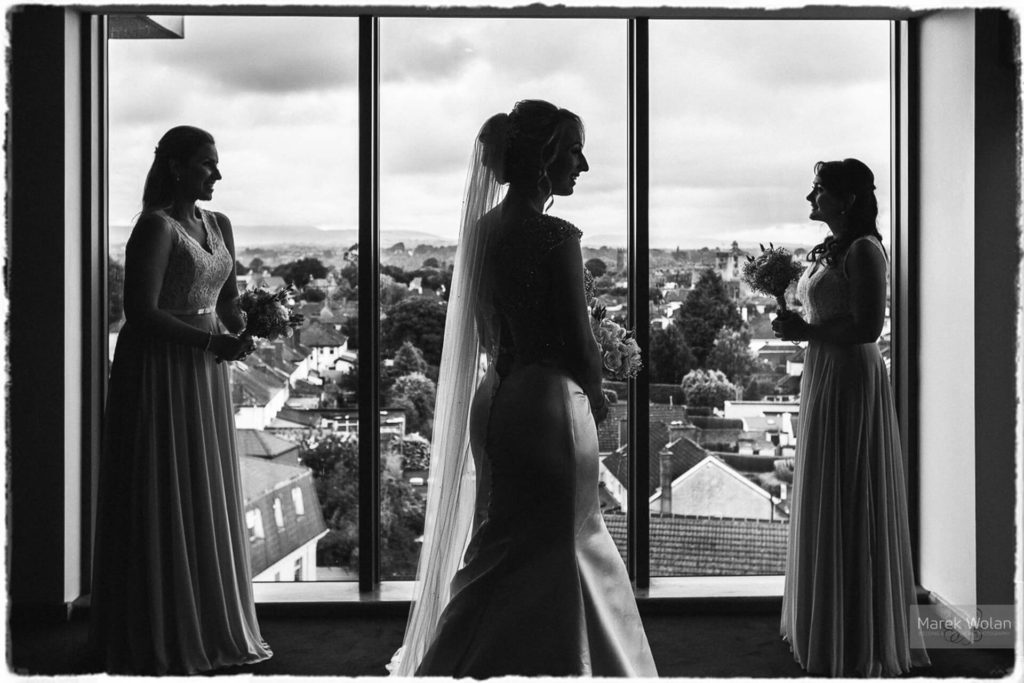 Three women in formal dresses, with one in a bridal gown and veil, stand by a large window overlooking a town, holding flower bouquets. The image is in black and white.