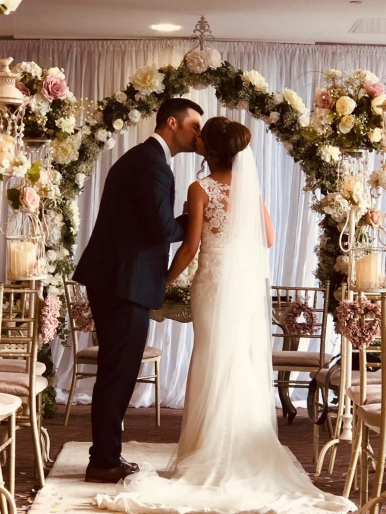 A bride and groom kiss under a floral wedding arch, holding hands and facing away from the seated guests inside a decorated venue.
