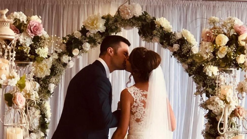 A bride and groom kiss in front of a floral arch decorated with white and pink flowers at their wedding ceremony.