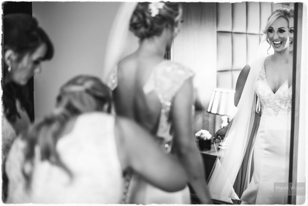 A bride in a wedding dress smiles at her reflection in a mirror while two women help her get ready.
