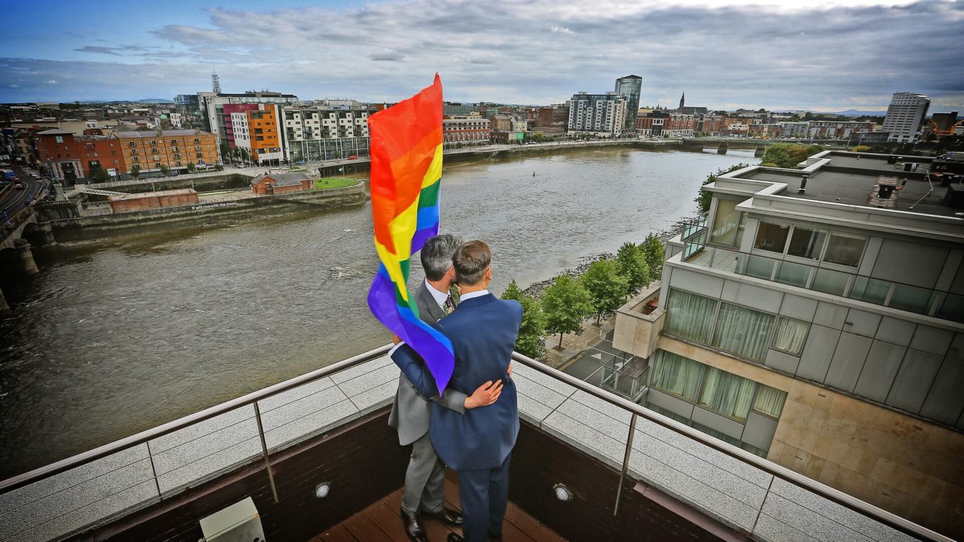 Two people embrace on a rooftop overlooking a river, holding a rainbow pride flag, with a cityscape in the background under a cloudy sky.