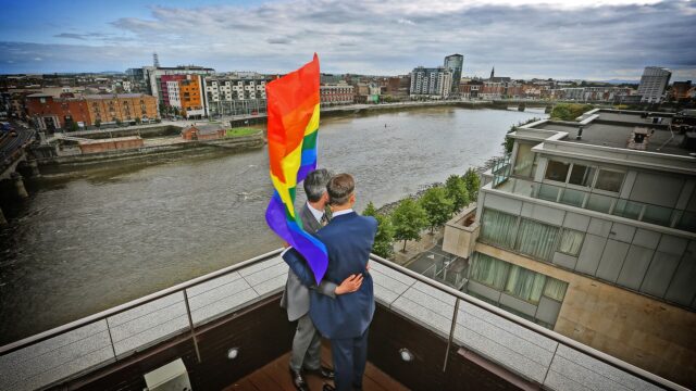 Two people embrace on a rooftop overlooking a river, holding a rainbow pride flag, with a cityscape in the background under a cloudy sky.