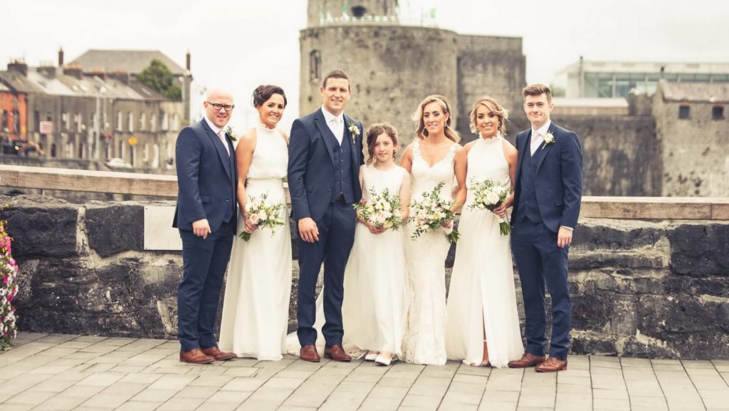 A wedding party of six adults and one child pose outdoors in formal attire, with stone walls and historic buildings in the background.