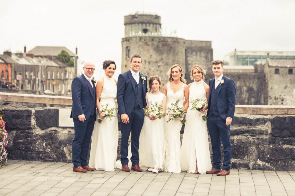 A wedding party of seven, dressed in formal attire, poses outdoors in front of a stone wall with a castle in the background.