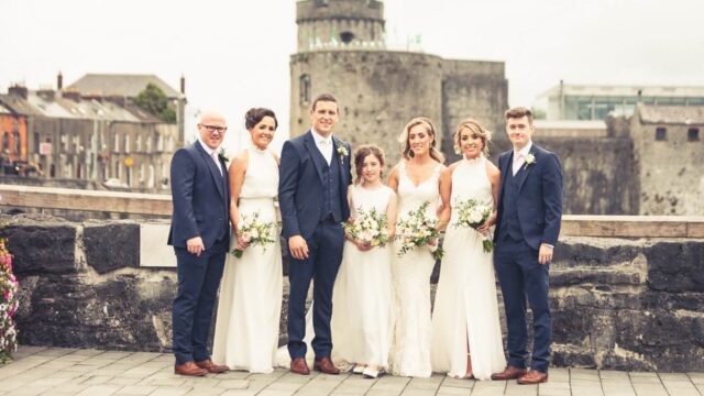 A wedding party of seven, dressed in formal attire, poses outdoors in front of a stone wall with a castle in the background.