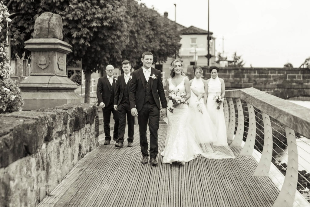 A bride and groom walk with their wedding party along a boardwalk, dressed in formal attire, with trees and stone structures in the background.
