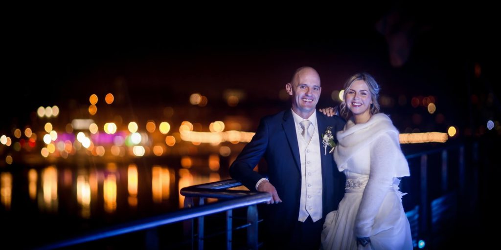 A bride and groom stand side by side on a railing at night, smiling, with city lights blurred in the background.