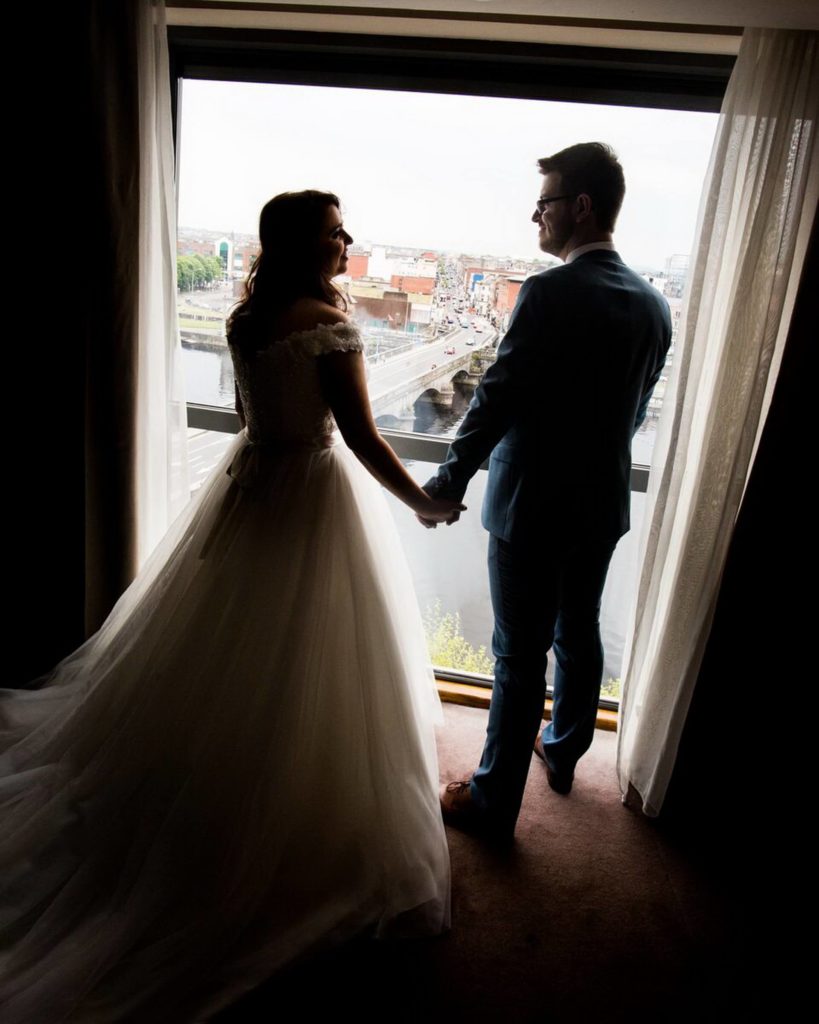A bride and groom, seen from behind, stand hand in hand by a window with city and river views, framed by sheer curtains.