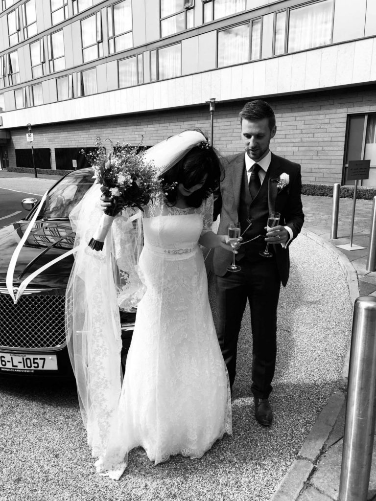 A bride adjusts her dress while holding a bouquet, standing beside a groom in a suit; both are outdoors near a car and building, holding champagne glasses.