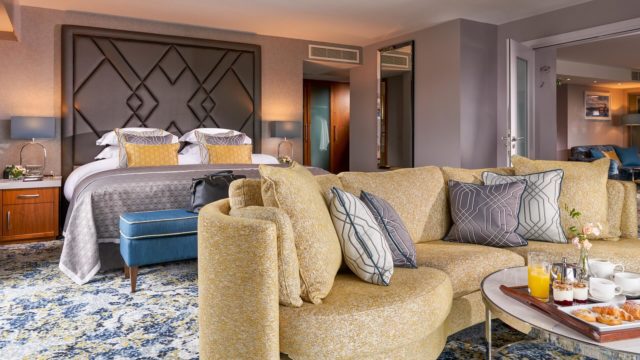 A modern hotel suite features a double bed, patterned sofa with pillows, ottoman, bedside tables with lamps, and a tray with drinks on a coffee table.