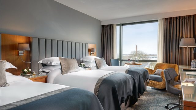 Modern hotel room with three single beds, grey and tan decor, a large window with curtains, armchair, desk, and view of trees outside.