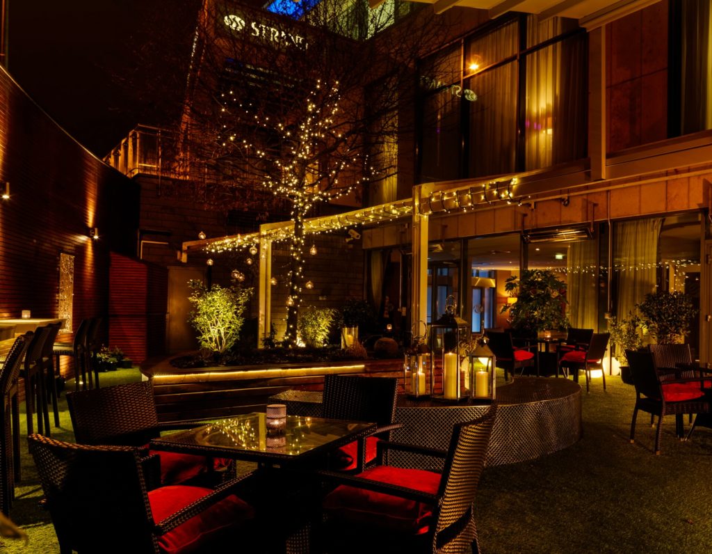 Outdoor courtyard of a restaurant or hotel at night, featuring string lights, illuminated trees, black wicker furniture with red cushions, and glass lanterns on tables.