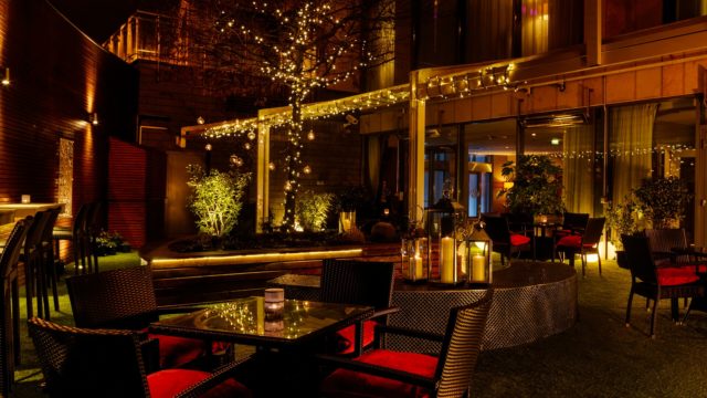 Outdoor patio at night with wicker chairs, glass tables, red cushions, string lights on trees, lanterns, and surrounding plants, creating a cozy and inviting atmosphere.