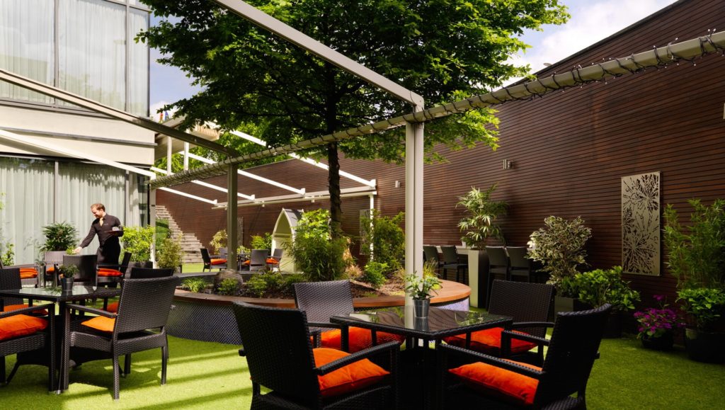 Outdoor dining area with black wicker tables and chairs, orange cushions, greenery, a central tree, and a person walking near the back.