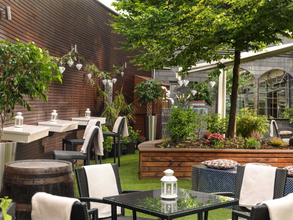Outdoor patio with glass tables, wicker chairs, potted plants, wooden walls, and greenery, featuring white lanterns and decorative cushions in a garden setting.
