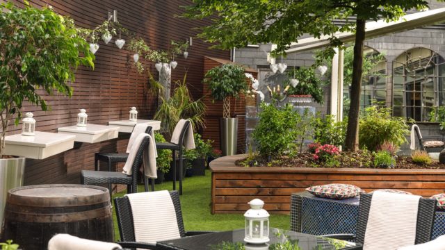 Outdoor patio with wicker chairs, wooden planters, greenery, small tables, and white lanterns; towels hang on chair backs and sinks are mounted on a wood-paneled wall.