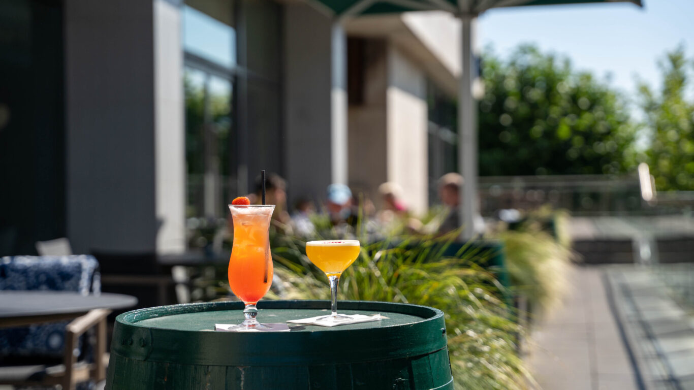 Two colorful cocktails sit on a green barrel table outside, with blurred people under umbrellas in the background—perfect for relaxing after exploring great Hotel Deals Limerick has to offer.