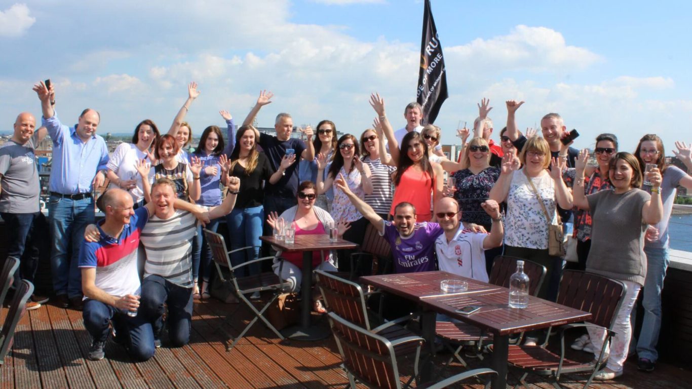 A group of people pose and wave for a photo on an outdoor deck with tables and chairs under a blue sky.