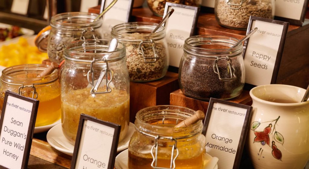 Jars of honey, marmalade, and poppy seeds with labeled signs are arranged on a wooden display at a breakfast buffet.