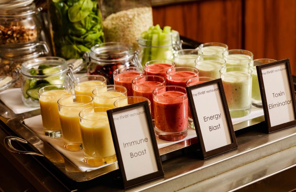 A tray with small glasses of yellow, red, and green smoothies labeled Immune Boost, Energy Blast, and Toxin Eliminator, with jars of ingredients in the background.
