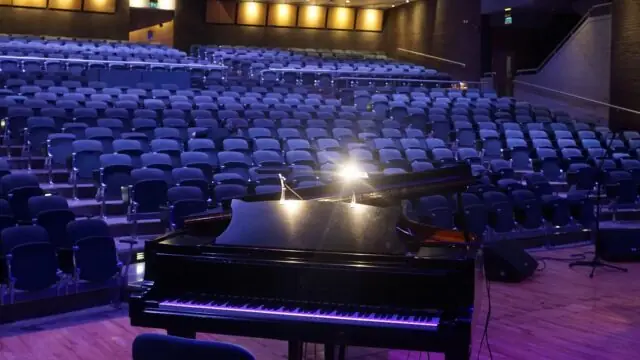 A grand piano on stage faces an empty auditorium with rows of blue seats and dim overhead lighting.