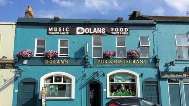A turquoise building with signs for “Dolan’s Pub & Restaurant,” advertising music seven nights a week and food seven days a week, with flowers on window sills and clear blue sky above.