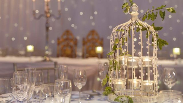Elegant table setting with empty wine glasses, a decorative birdcage centerpiece holding candles, and a candelabra in the background against a softly lit backdrop.