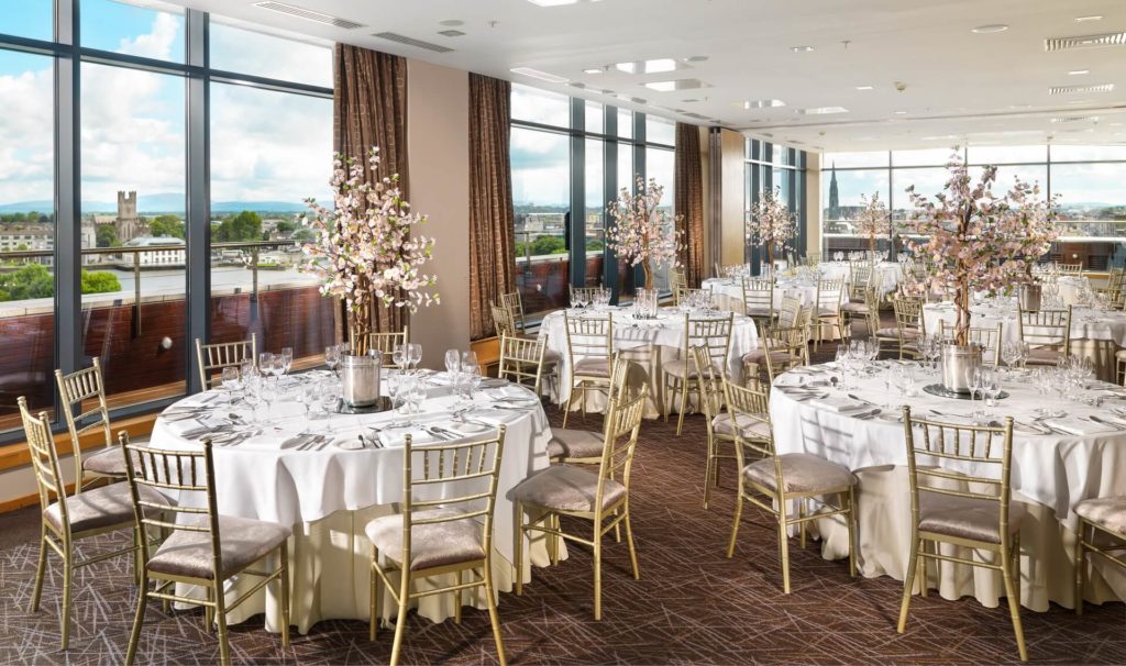 Elegant banquet hall with round tables set for an event, featuring white tablecloths, gold chairs, and floral centerpieces, with large windows overlooking a cityscape.