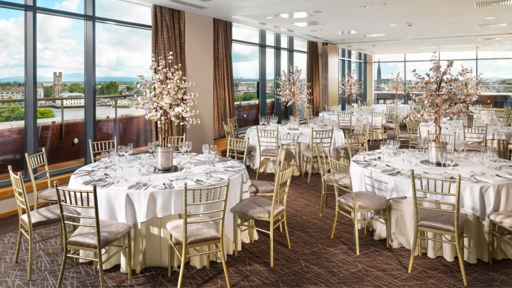 Banquet hall with round tables covered in white cloths, gold chairs, formal table settings, and tall floral centerpieces near large windows with a city view.