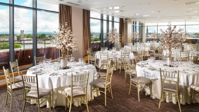 A banquet hall set for an event with round tables, white tablecloths, gold chairs, floral centerpieces, and large windows overlooking a cityscape.