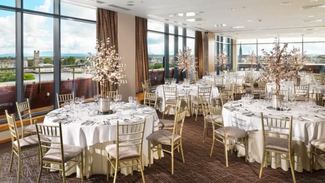 Elegant banquet hall with floor-to-ceiling windows, round tables set for dining, gold chairs, and decorative floral centerpieces. City view visible through large windows.