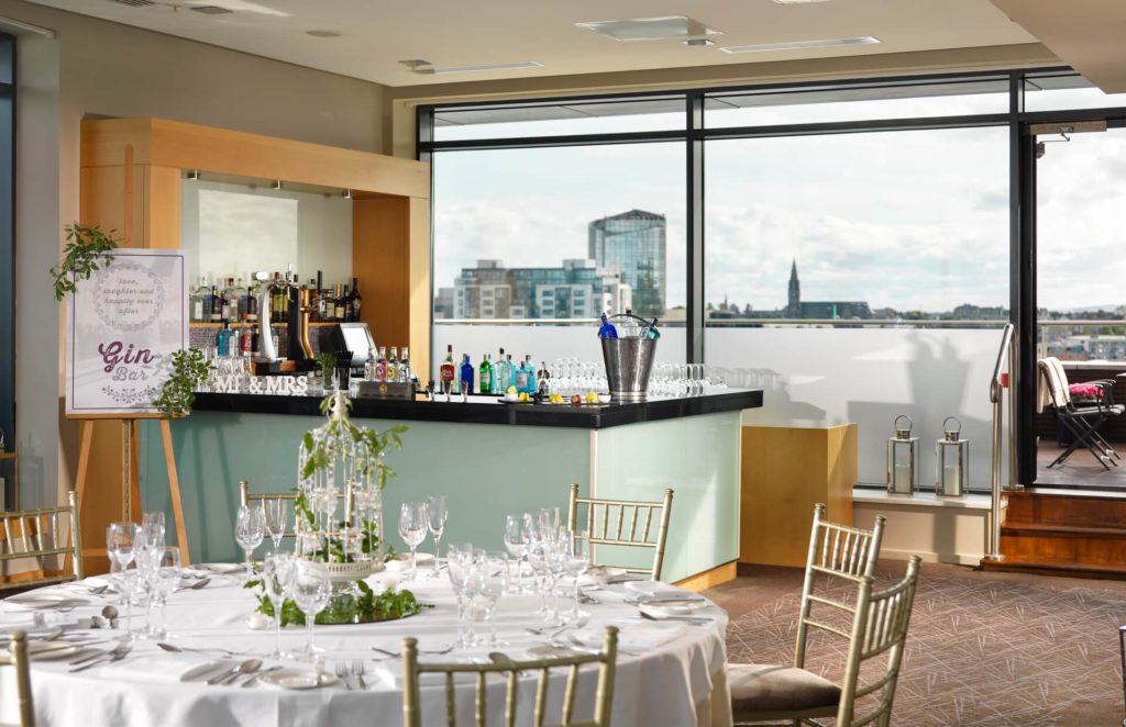 A modern event space with a set dining table, decorated gin bar, and large windows overlooking a city skyline in daylight.