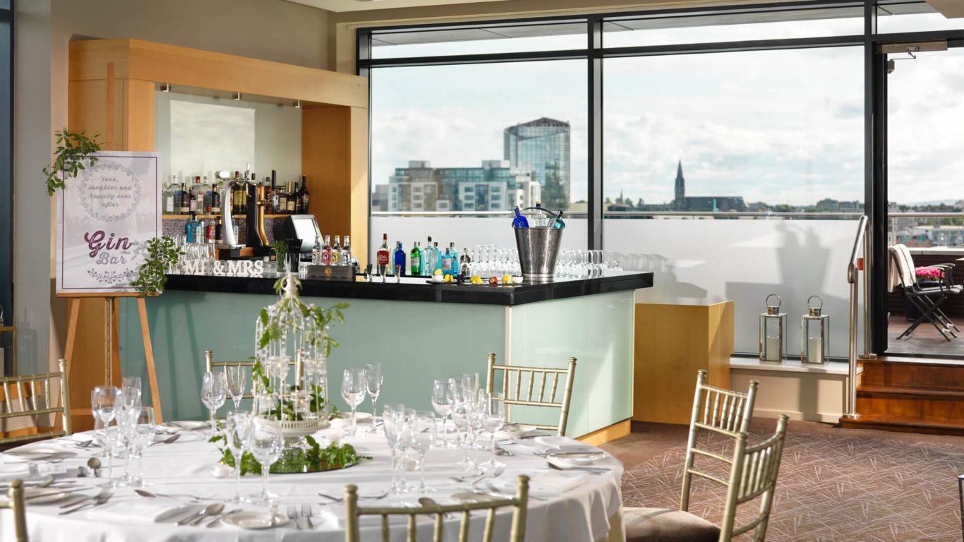 Indoor wedding reception setup with a round table set for guests, a gin bar in the background, and large windows revealing a cityscape view.