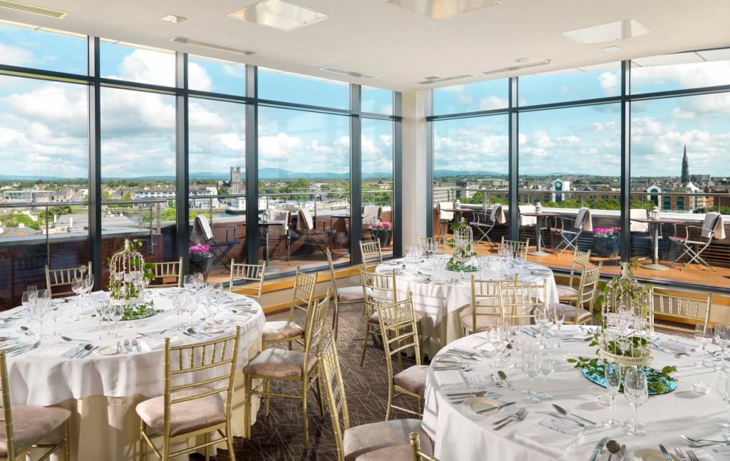 A bright banquet room with round tables set for dining, gold chairs, and floor-to-ceiling windows offering a cityscape view under a partly cloudy sky.